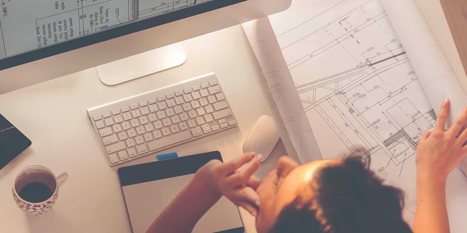 Architect reviewing blueprints at a desk with computer and drawing tablet.
