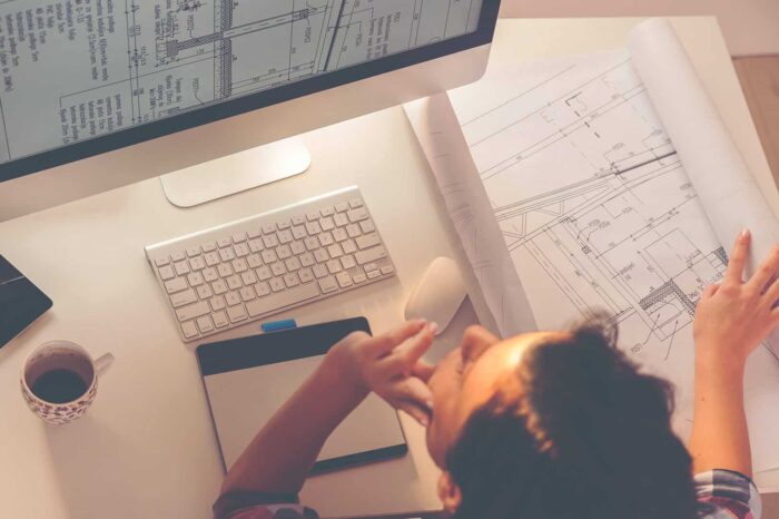 Architect reviewing blueprints at a desk with computer and drawing tablet.
