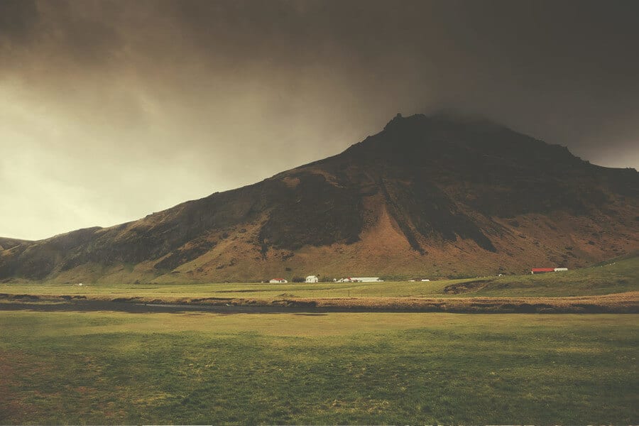 Icelandic landscape with a large mountain under a cloudy sky. Green field in foreground.